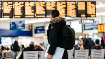 man standing inside airport looking at LED flight schedule bulletin board
