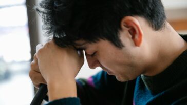 Close-up of a man looking stressed as he rests his head on his arms indoors.