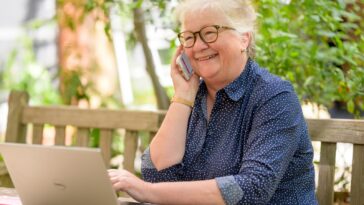 a woman on her phone while sitting at a table with a laptop