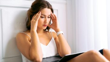 Young woman sitting with hands on head, looking stressed while working on a laptop indoors.
