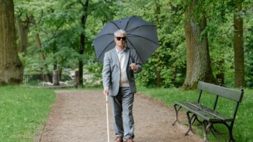 Senior man with visual impairment walks in a park with a walking stick and umbrella.