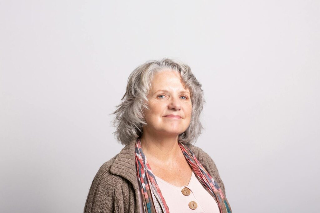 A cheerful elderly woman with gray hair smiling warmly in a neutral setting.