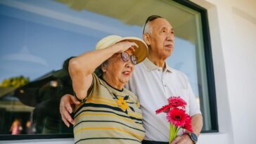 Senior couple standing outside, holding flowers, enjoying a sunny day together.