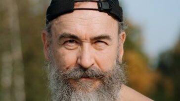 A senior man with a beard and cap poses shirtless outdoors, exuding confidence.