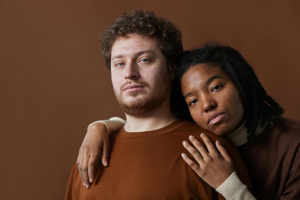 A couple with diverse ethnicities embracing with a serious expression on a brown backdrop.