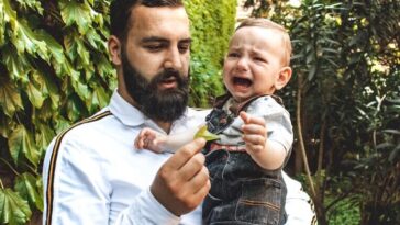 A bearded father comforts his crying baby while holding him in a lush green garden.