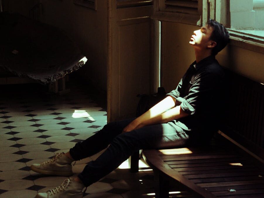 A young man sits in a dimly lit room, soaked in sunlight, depicting introspection.