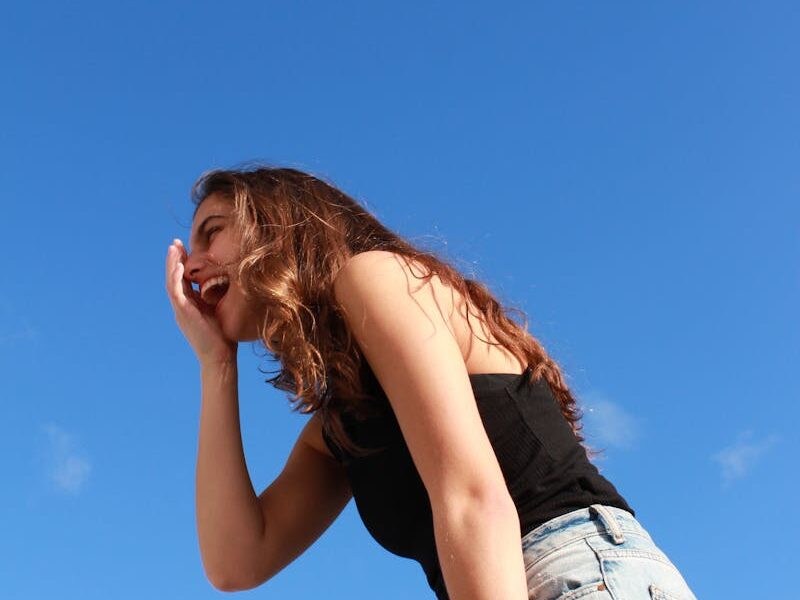 A young woman in casual attire laughing outdoors against a vibrant blue sky, radiating happiness.