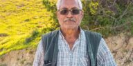 Portrait of an elderly man standing outdoors on a sunny day, in a rural setting.