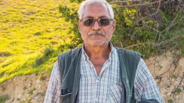 Portrait of an elderly man standing outdoors on a sunny day, in a rural setting.