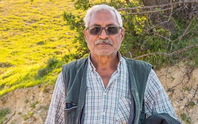 Portrait of an elderly man standing outdoors on a sunny day, in a rural setting.