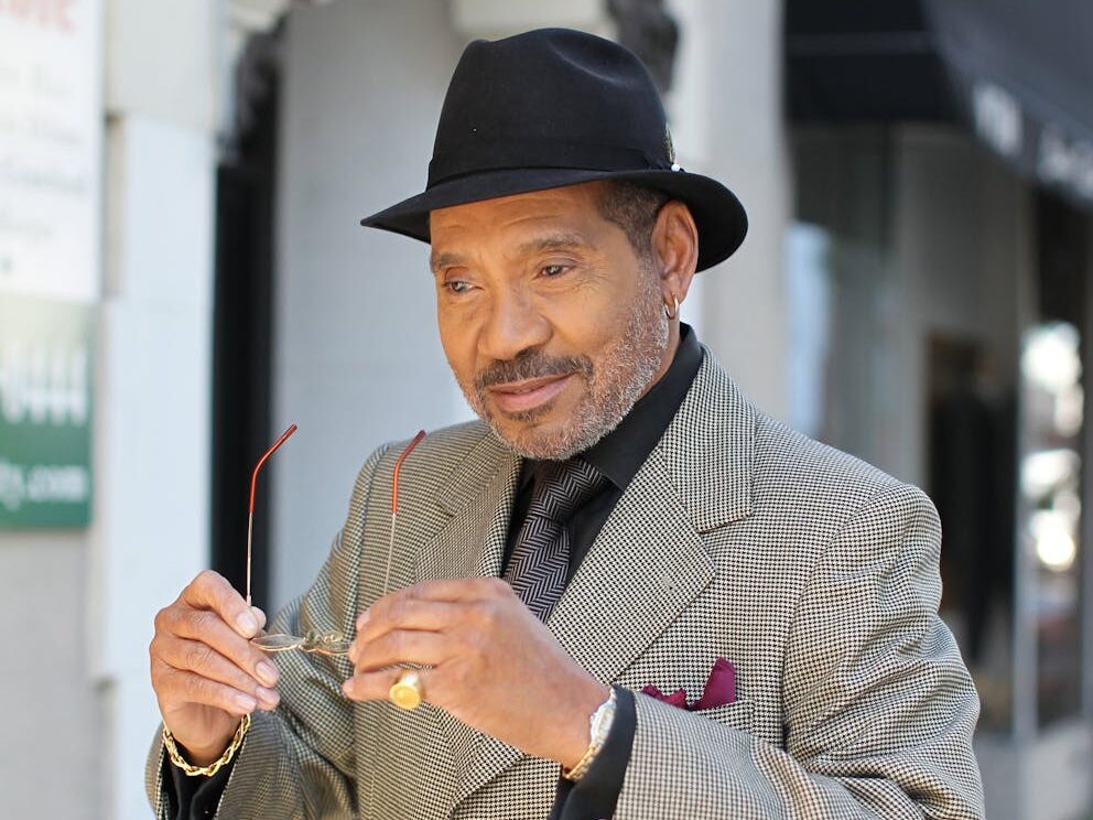 An elegant senior man in a formal coat and fedora smiling outdoors, adjusting his eyeglasses.