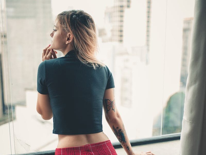 A thoughtful woman stands by a window, overlooking the cityscape in casual attire.