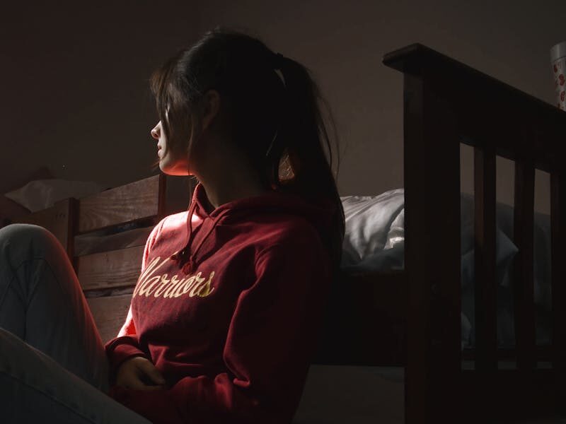 Contemplative young woman in hoodie sitting on the floor in dimly lit room, symbolizing solitude and introspection.