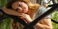 A serene portrait of a woman with long hair leaning on a metal structure in a park. Sun-dappled and peaceful.