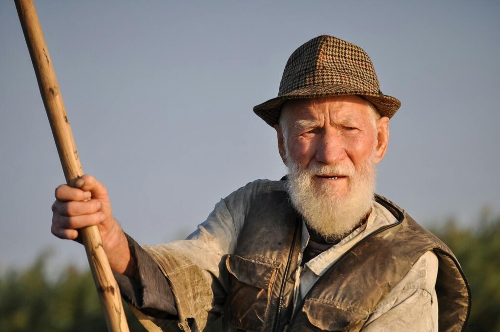 Portrait of a senior fisherman holding a rod, wearing a hat and vest outdoors.