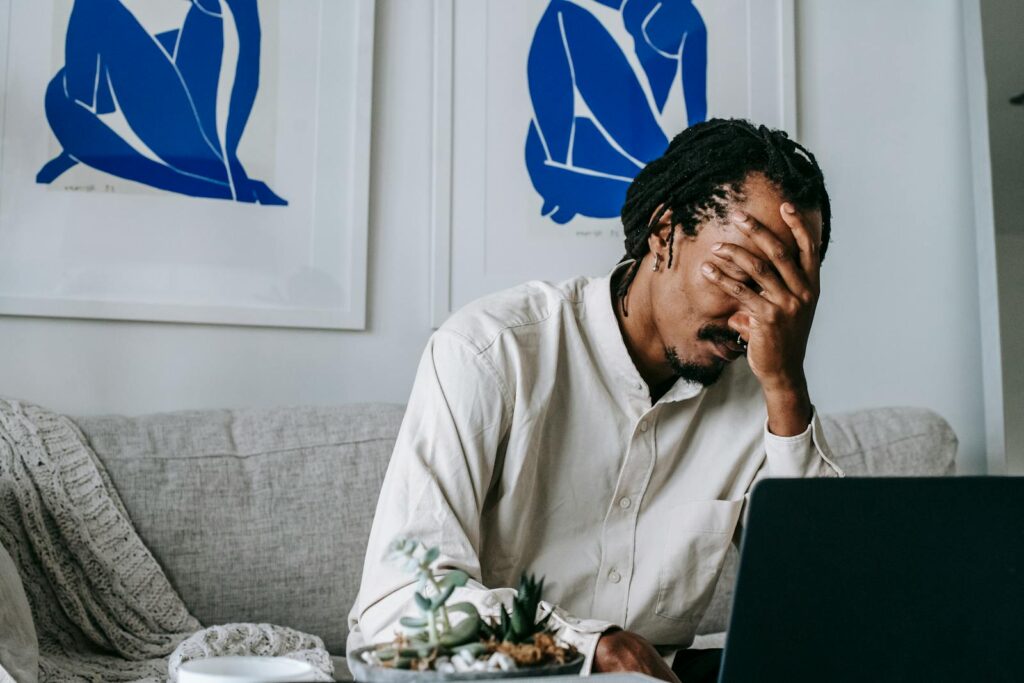 Frustrated young bearded African American male freelancer with dreadlocks in casual shirt working on laptop at home and covering eyes with hand after failure