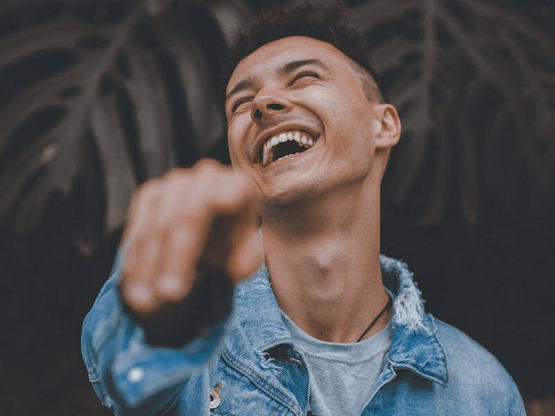 A joyful man in a denim jacket laughs and points, surrounded by lush greenery.