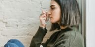 Side view of depressed young ethnic female millennial in warm sweater sitting leaning on wall and wiping tears with tissue at home