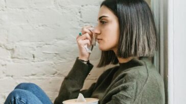 Side view of depressed young ethnic female millennial in warm sweater sitting leaning on wall and wiping tears with tissue at home