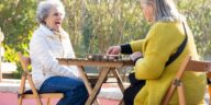 Two senior women laughing and playing dominoes outdoors on a sunny day.
