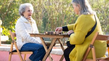 Two senior women laughing and playing dominoes outdoors on a sunny day.