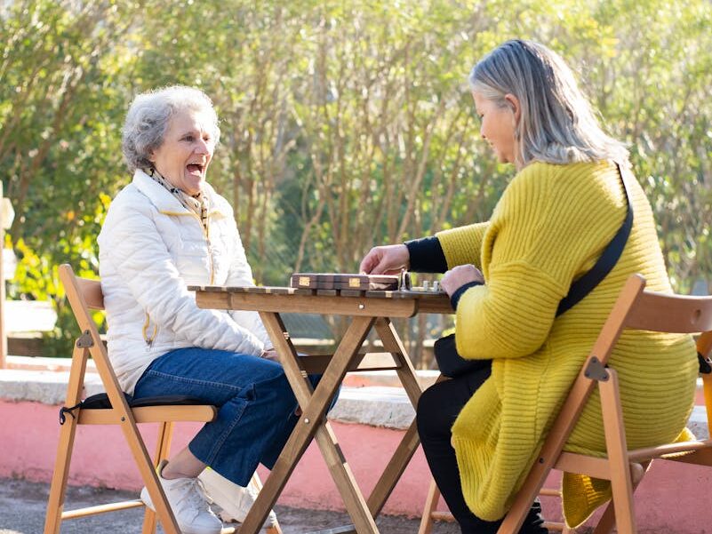 Two senior women laughing and playing dominoes outdoors on a sunny day.
