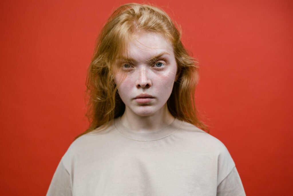 A young woman with ginger hair looking intently at the camera against a vibrant red backdrop.