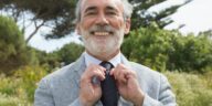 Senior man in suit smiling while adjusting his necktie outdoors against a lush green background.