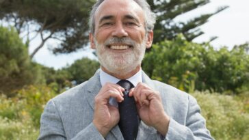 Senior man in suit smiling while adjusting his necktie outdoors against a lush green background.